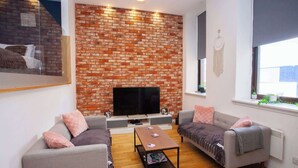 A modern living room with a brick wall, a flat-screen TV, two grey sofas, a coffee table, and a window with plants. - Stunning City Centre Duplex (Glasgow City)