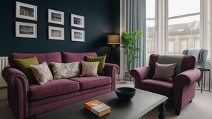 Una sala de estar con pared azul oscuro, un sofá morado, un sillón gris y una mesa de centro. Hay cuadros enmarcados en la pared y una lámpara en la esquina.