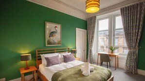 Habitación de hotel con pared verde, cama con cubrecama verde, cabecera de madera, mesita de noche con lámpara, escritorio con silla y ventana con cortinas.