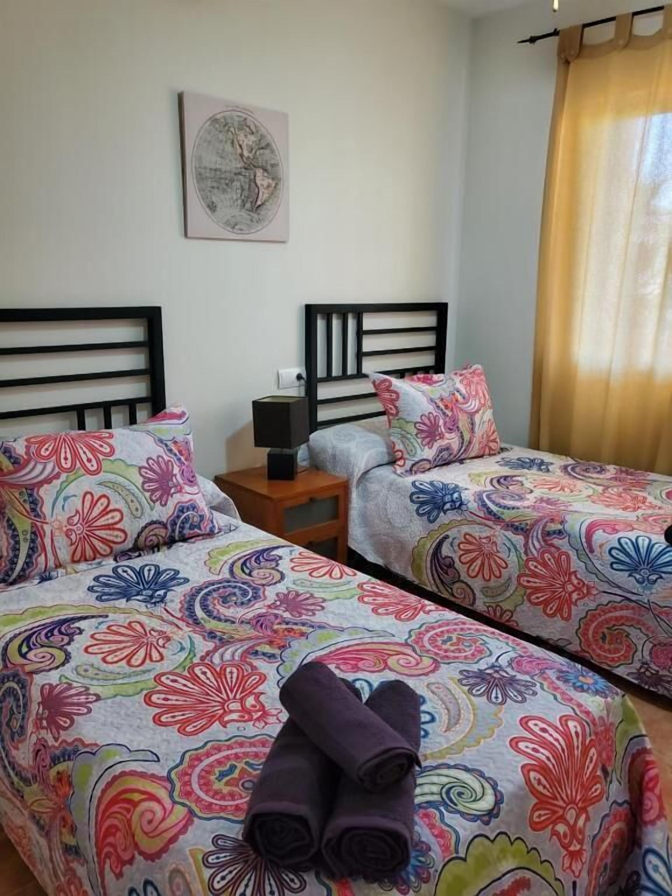 Habitación de hotel con dos camas, cada una con una colcha floral de colores. Hay una mesita de noche con una lámpara y una pequeña mesa entre las camas. Un cuadro enmarcado cuelga de la pared sobre las camas.