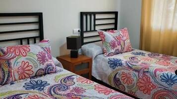 Habitación de hotel con dos camas, cada una con una colcha floral de colores. Hay una mesita de noche con una lámpara y una pequeña mesa entre las camas. Un cuadro enmarcado cuelga de la pared sobre las camas.