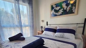 A neatly made bed with floral bedding, a painting of daisies on the wall, and a window with curtains.