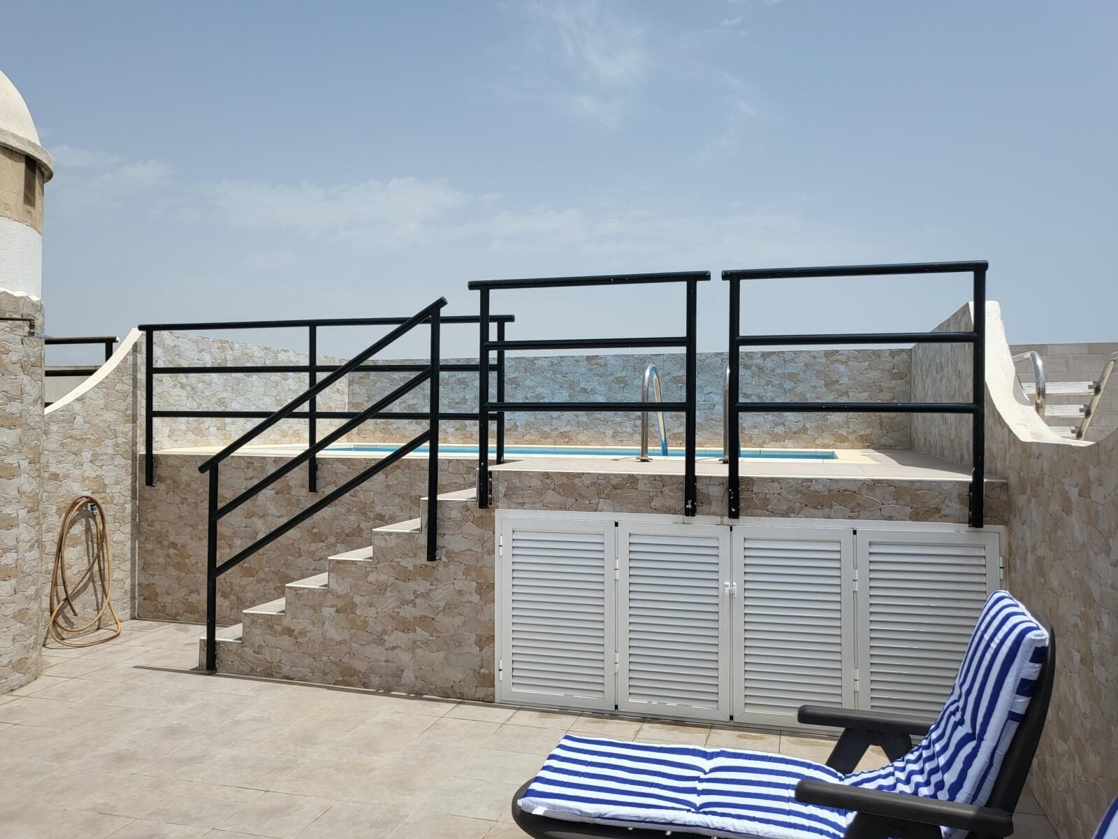Un área de piscina en la azotea con un muro de piedra, una escalera de piscina y una silla de descanso con cojín a rayas.