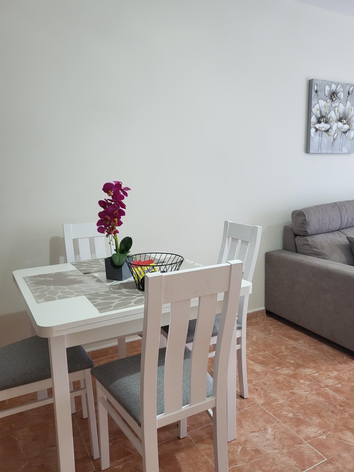 Un coin repas avec une table et des chaises blanches, un vase rempli de fleurs violettes et un tableau accroché au mur.