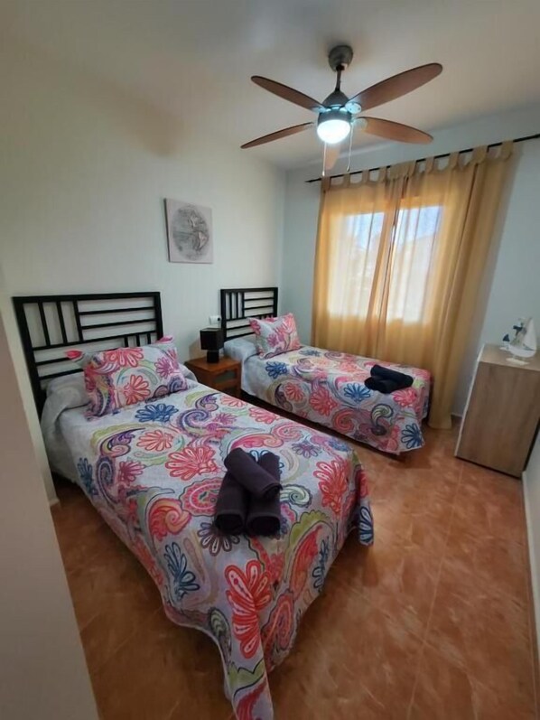 Une chambre avec deux lits, un ventilateur de plafond, une table de chevet et une fenêtre avec des rideaux.