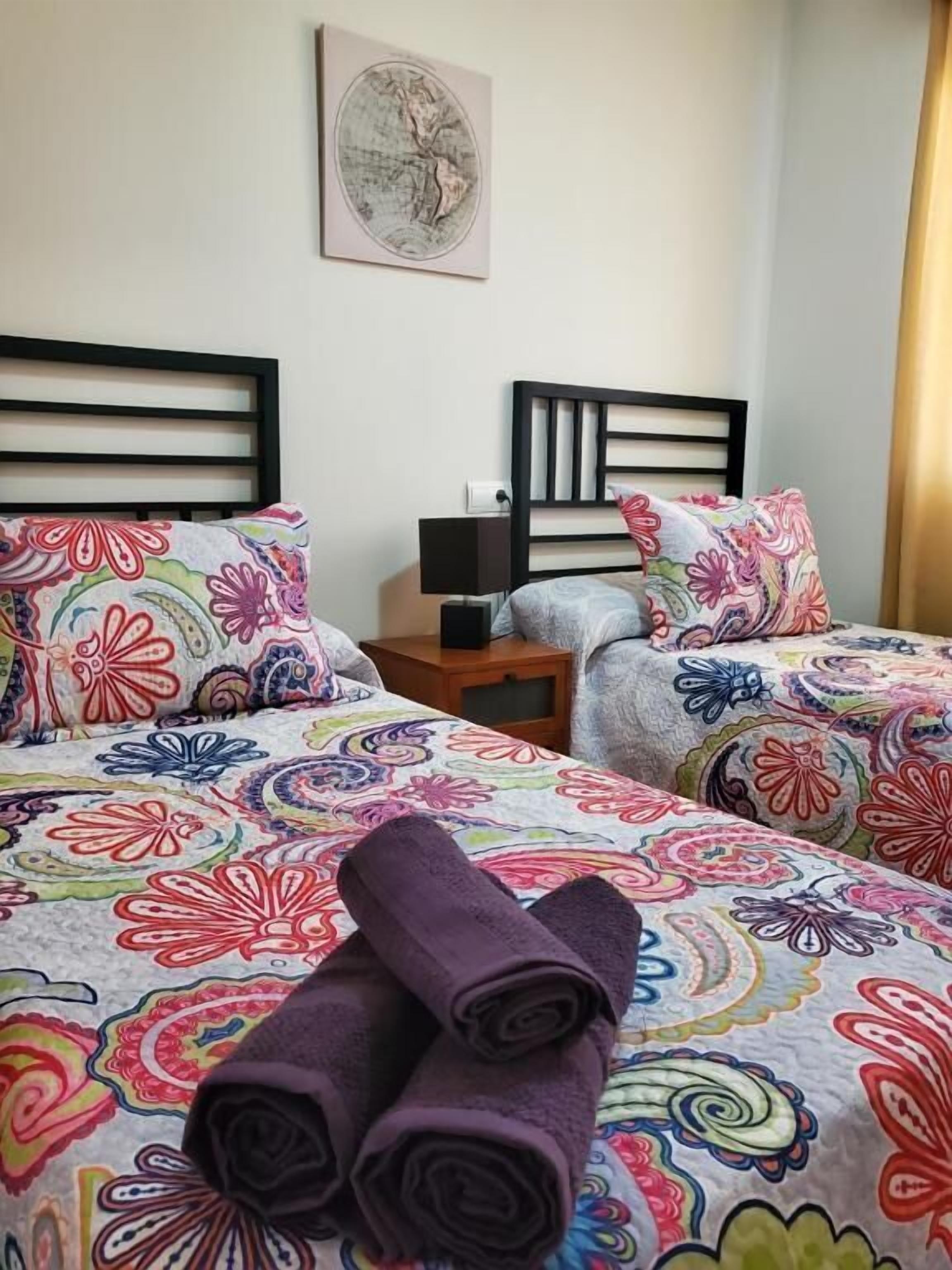 A hotel room with two beds, colorful floral bedding, a nightstand with a lamp, and a wall-mounted painting.