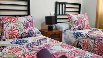 A hotel room with two beds, colorful floral bedding, a nightstand with a lamp, and a wall-mounted painting.