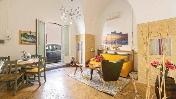 Ein gemütliches Wohnzimmer mit Essbereich, einer Couch und einem Holztisch. Eine Tür führt nach draußen, ein Gemälde ist an der Wand und eine Chandeliebre leuchtet.