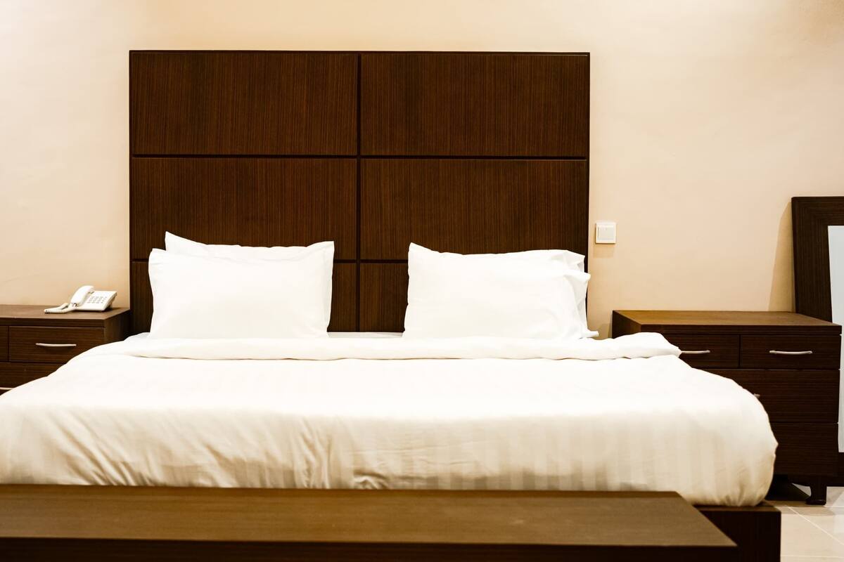 A hotel room with a large bed, two bedside tables, a nightstand, a wooden headboard, and a bench.
