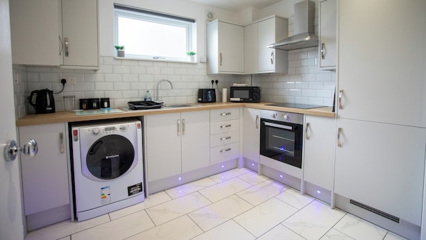 Una cocina moderna con muebles blancos, horno empotrado, microondas, lavadora y una ventana con una planta.