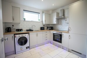 A modern kitchen with white cabinetry, a built-in oven, a microwave, a washing machine, and a window with a plant. - 3 bed Apartment in Huddersfield (Huddersfield)
