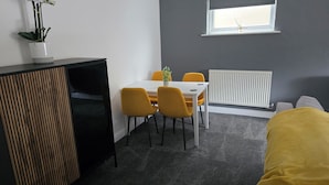 Una habitación pequeña con una mesa blanca, cuatro sillas amarillas, un armario negro, un radiador y una ventana con una planta en el alféizar.