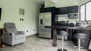 A modern kitchen with a stainless steel refrigerator, a microwave, and a stove. There is a grey armchair and two bar stools. - Palo Rosa 1 (Quepos)
