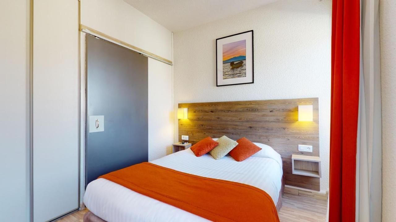 Une chambre d’hôtel comprenant un lit, une tête de lit en bois, deux tables de chevet, une petite table et une lampe fixée au mur.