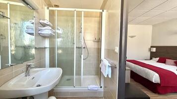 A hotel bathroom with a shower, sink, and a bed with a red blanket.