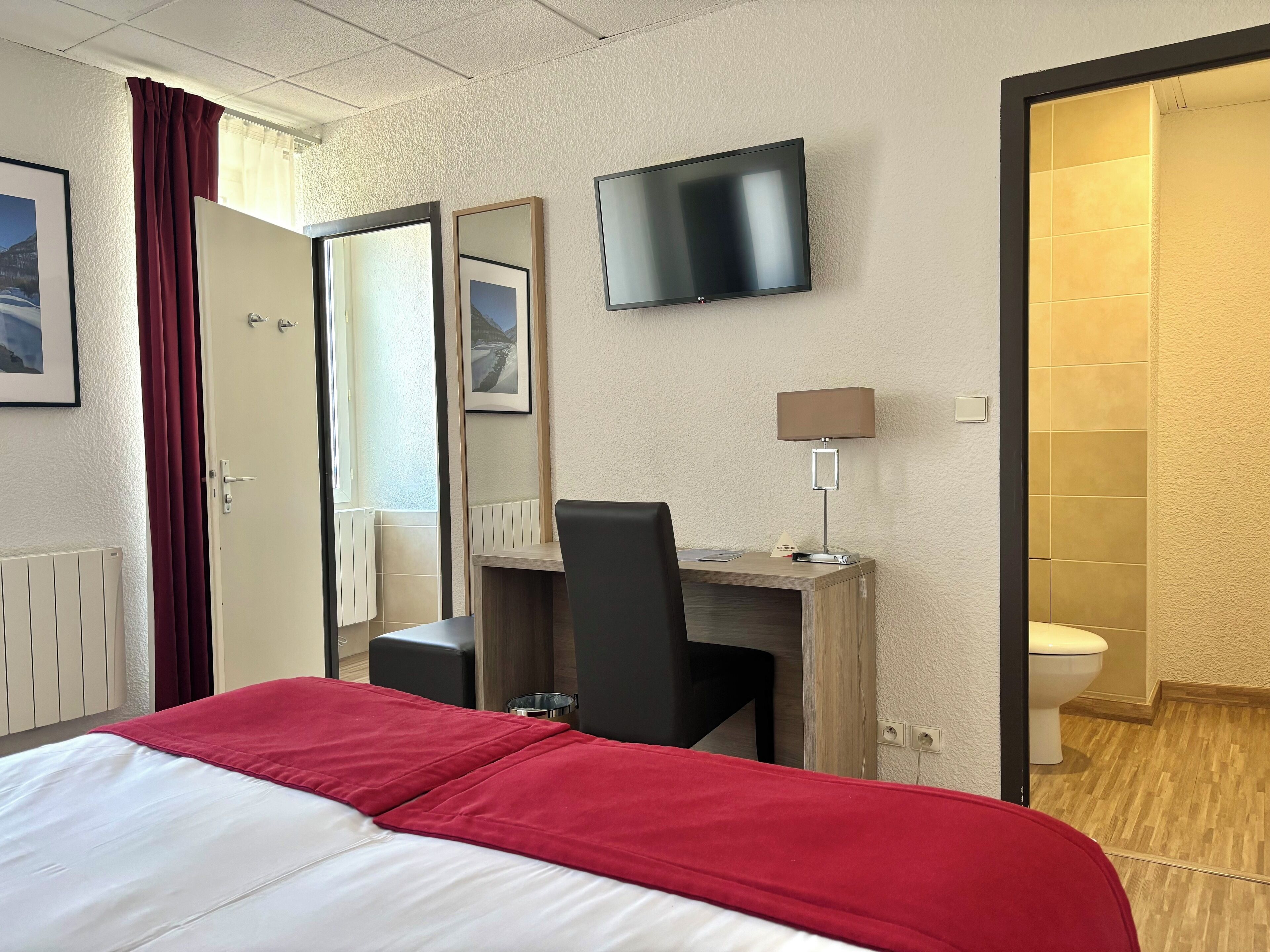 Habitación de hotel con una cama, un escritorio con silla, televisor, espejo y baño con inodoro.