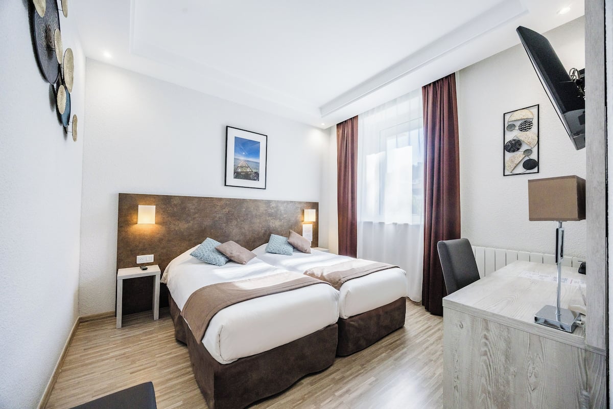 A hotel room with a double bed, bedside tables, a desk, a chair, a lamp, and a view of the city.