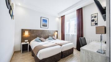A hotel room with a double bed, bedside tables, a desk, a chair, a lamp, and a view of the city.