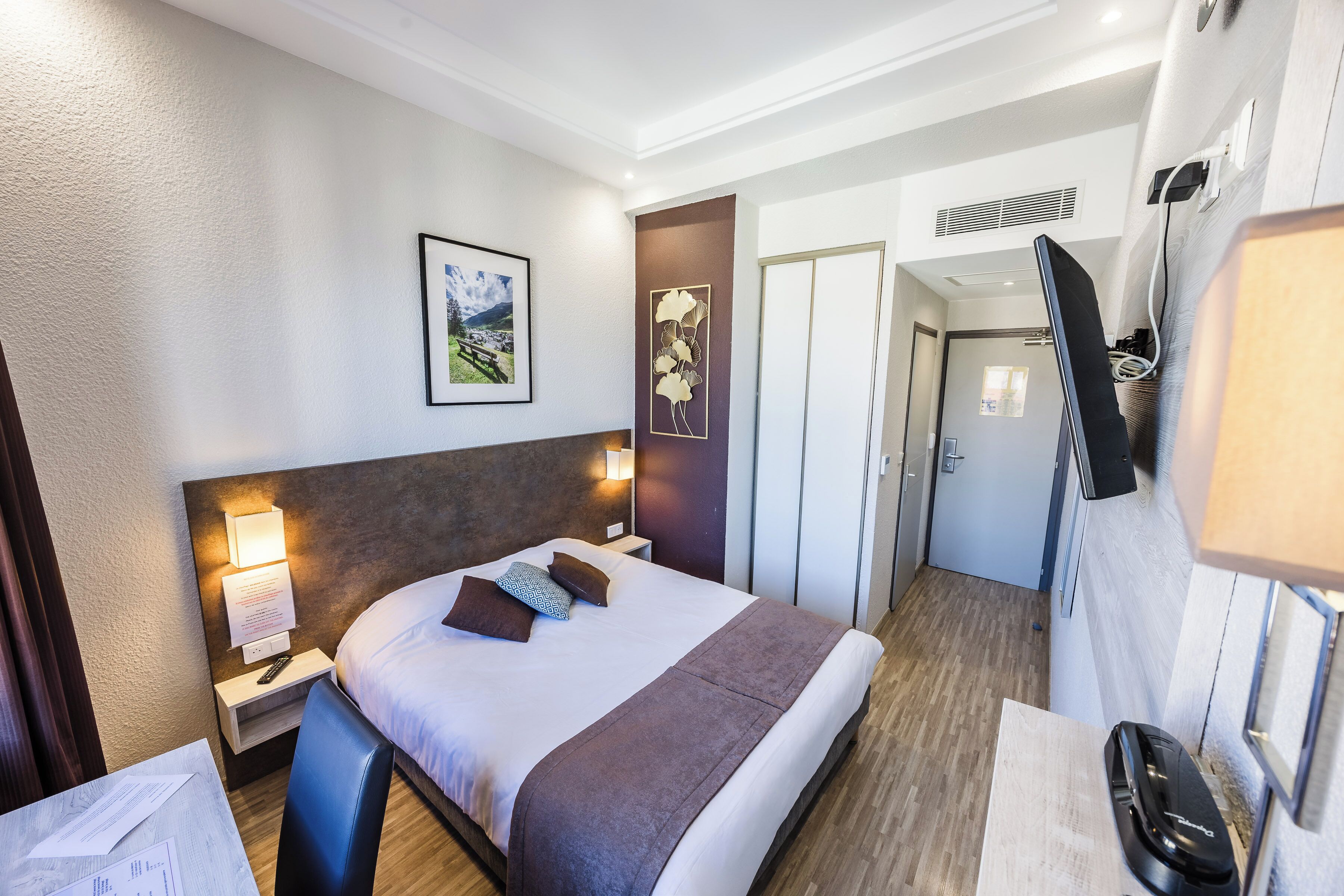 Habitación de hotel con cama, mesitas de noche, televisor, un cuadro enmarcado en la pared y una puerta que da a otra habitación.