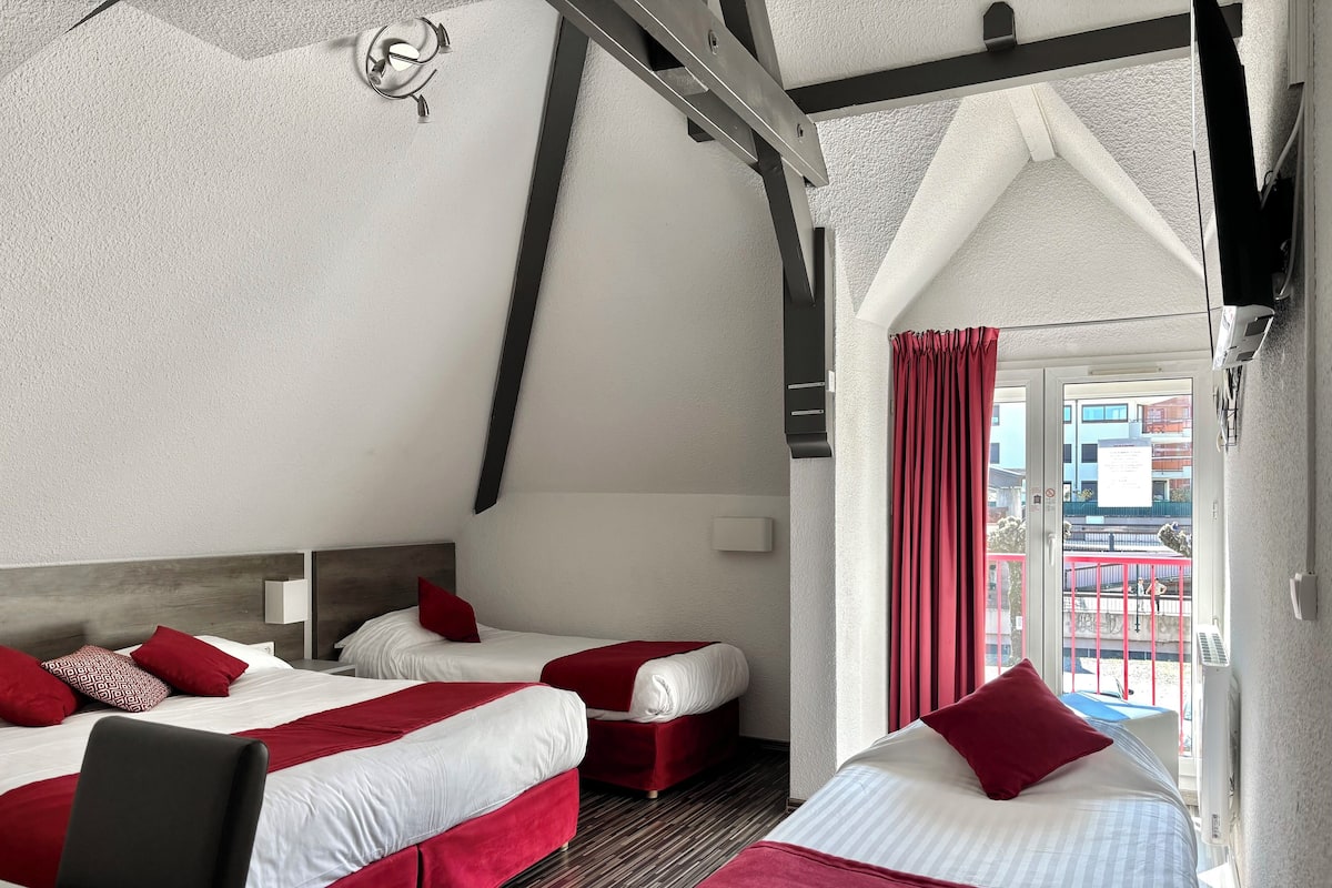 A hotel room with two beds, red and white bedding, a wooden headboard, a small bedside table, and a window with red curtains.