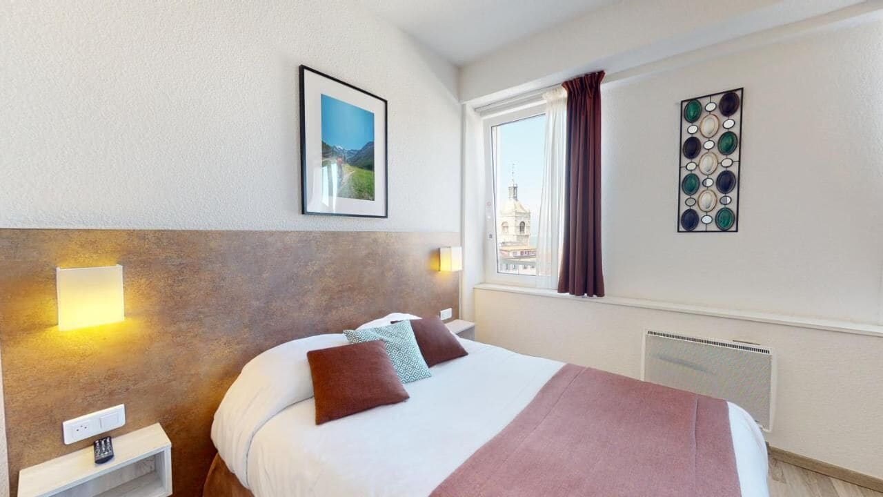 A hotel room with a bed, bedside lamps, a window with curtains, a wall-mounted picture, and a radiator.