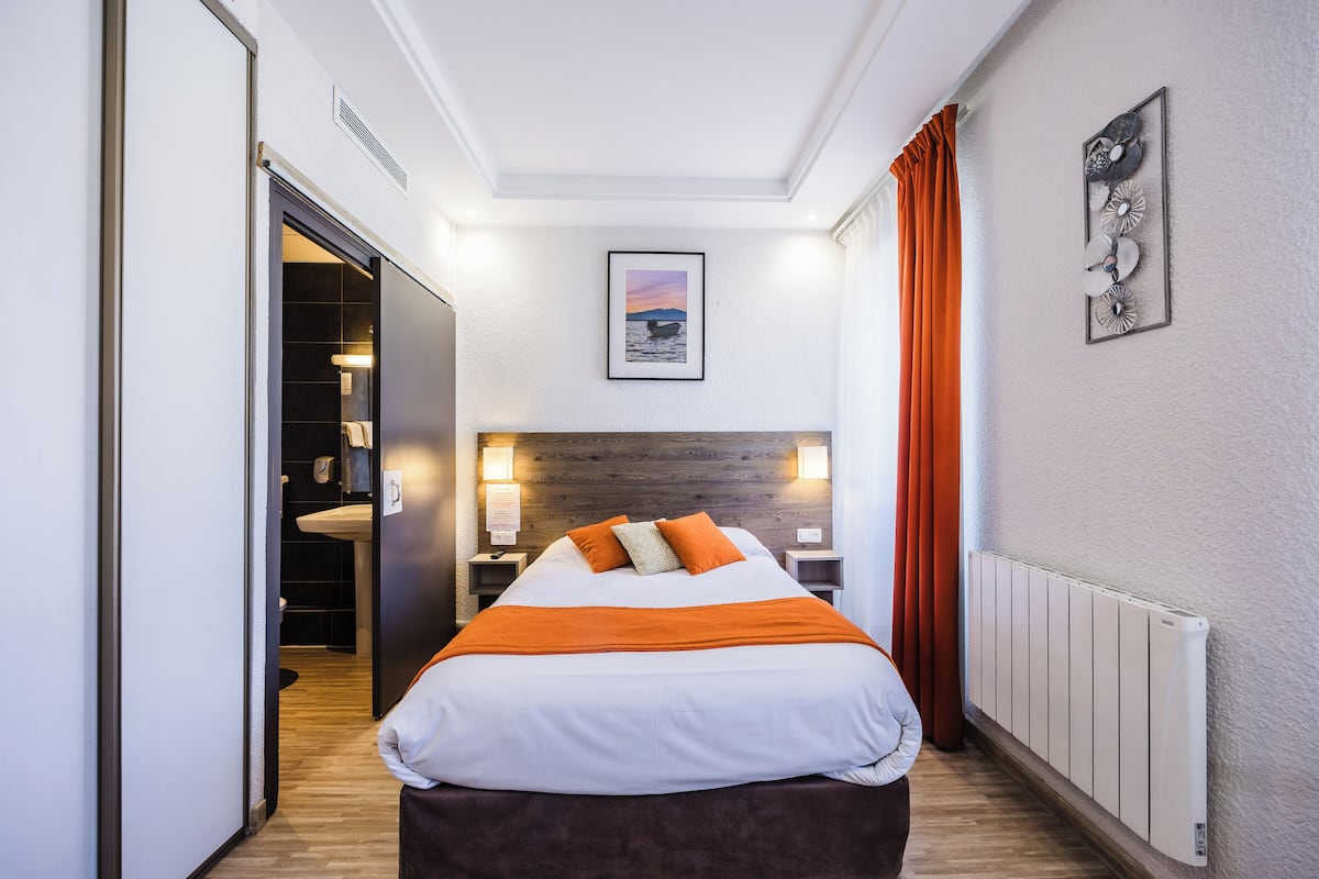 A hotel room with a bed, a nightstand, a wall-mounted light, a radiator, and a door leading to a bathroom.