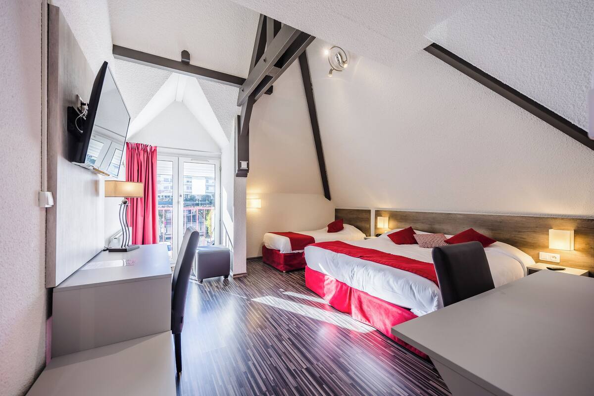 A hotel room with a sloped ceiling, two beds with red and white bedding, a desk with a chair, and a view of the city through a window.