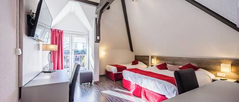A hotel room with a sloped ceiling, two beds with red and white bedding, a desk with a chair, and a view of the city through a window.
