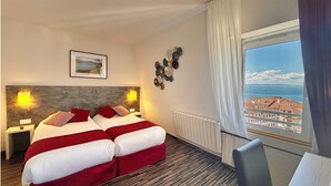 Habitación de hotel con una cama, dos mesitas de noche con lámparas, un cuadro en la pared, decoración mural, una ventana con vistas a edificios y al mar, y un radiador.