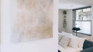 A modern living room with a large abstract painting on the wall, a sofa with patterned cushions, and a window with blinds. - Designer Flat in Heart of Brighton - Walk to Beach & Attractions! (Brighton and Hove)