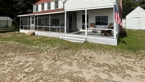 A white two-story house with a red roof, a porch with outdoor seating, and an American flag. - LARGE RUSTIC COTTAGE. SEBAGO LAKE BEACHFRONT (SEBAGO)
