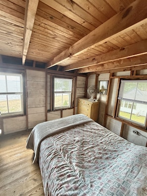 A bedroom with a wooden ceiling, a bed with a patterned bedspread, a dresser, and two windows with blinds. - LARGE RUSTIC COTTAGE. SEBAGO LAKE BEACHFRONT (SEBAGO)