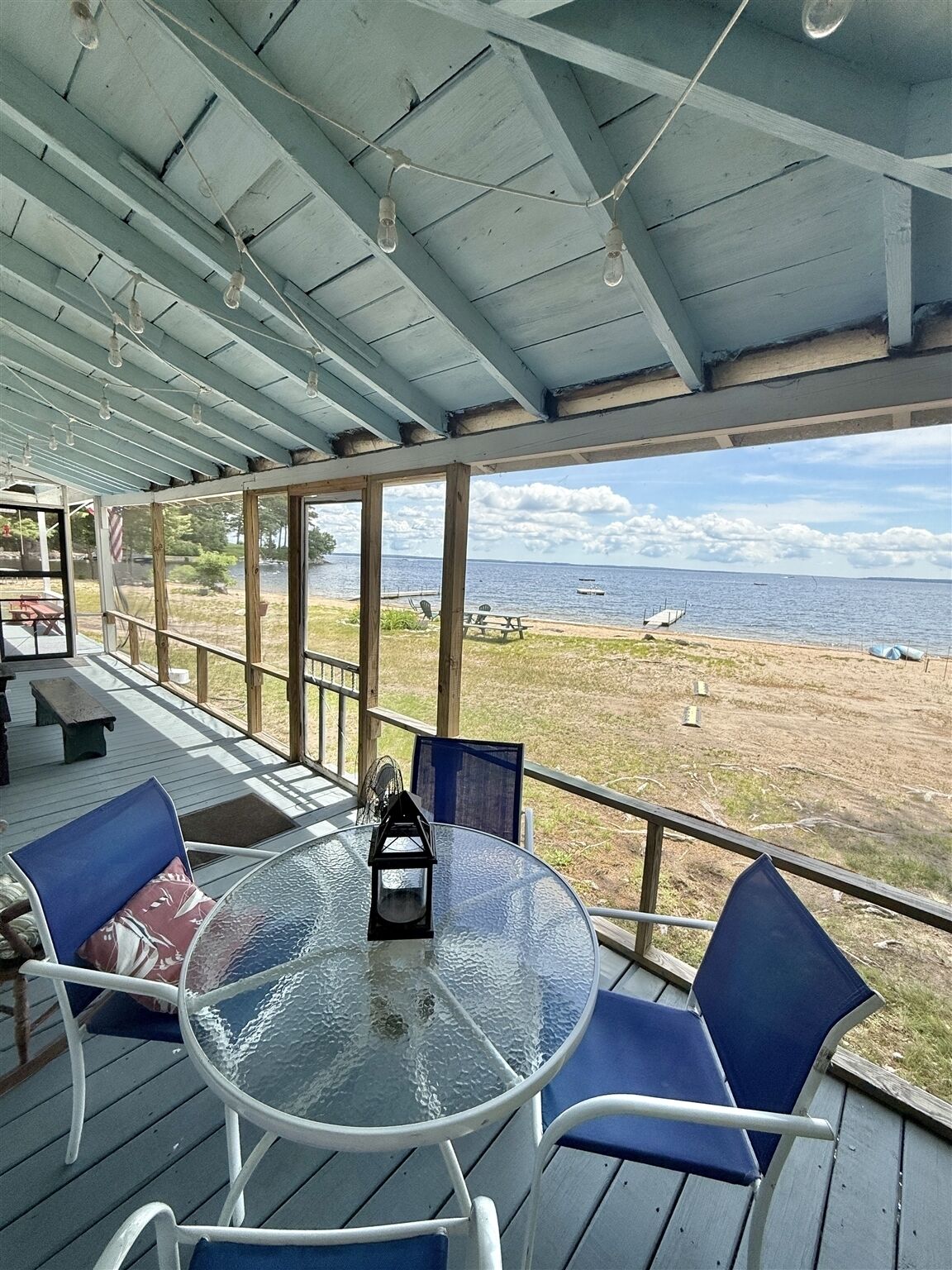 A covered outdoor seating area with a glass table and chairs, overlooking a beach and the sea.
