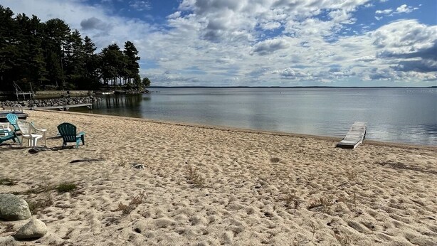 A beach with a sandy shore, a few lounge chairs, and a calm body of water. - LARGE RUSTIC COTTAGE. SEBAGO LAKE BEACHFRONT (SEBAGO)