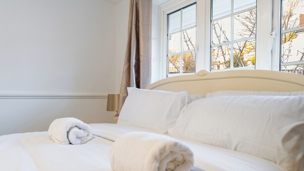Una cama bien tendida con sábanas blancas y dos toallas enrolladas, una ventana con vista a árboles desnudos y un cabecero color beige.