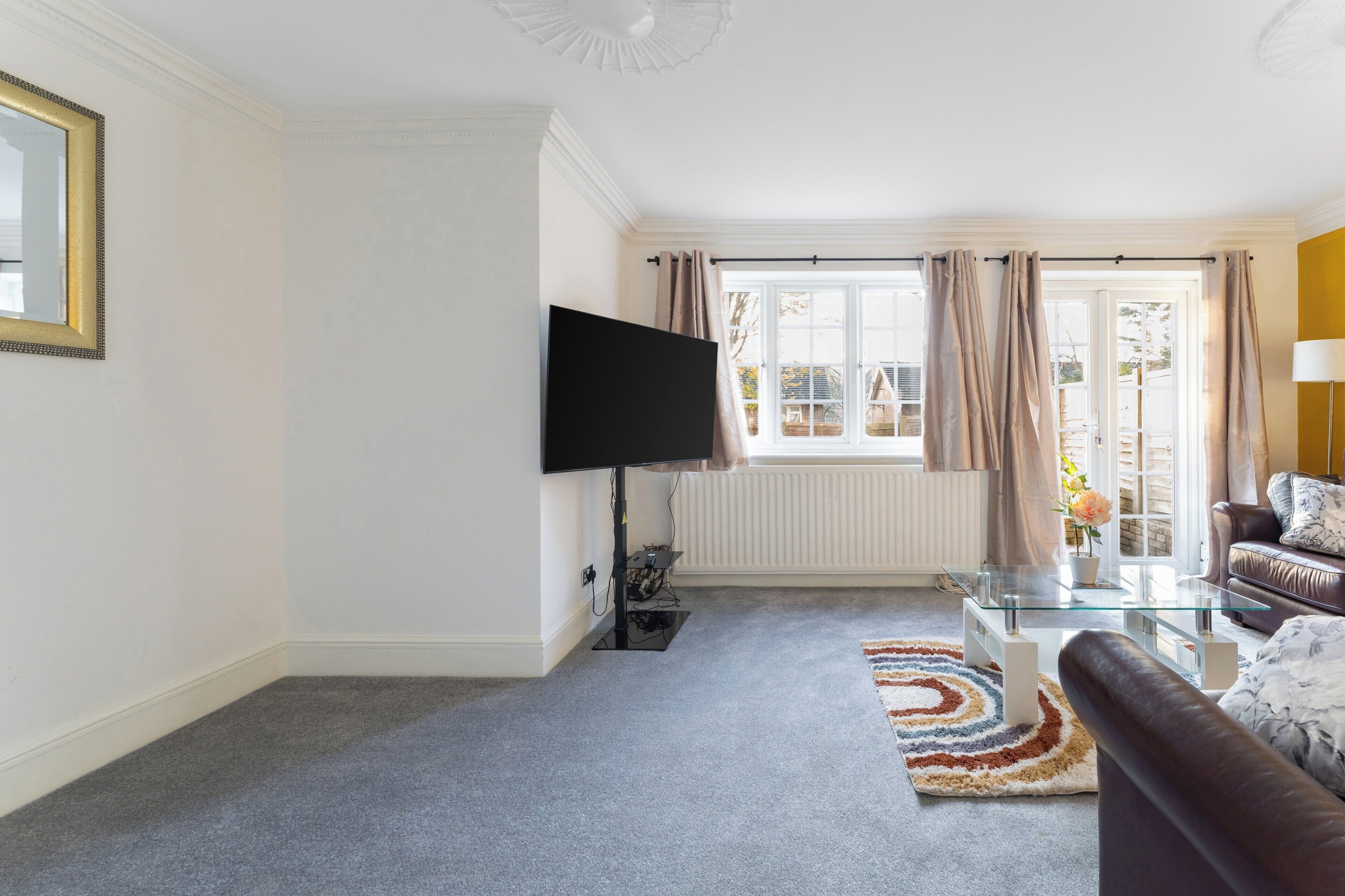 A living room with a flat-screen TV, a glass coffee table, a brown leather sofa, and a patterned rug.