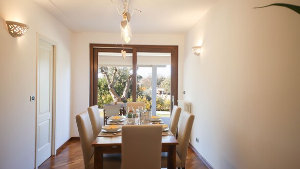 A dining area with a wooden table set for four, surrounded by chairs, with a view of trees and a building outside through large windows. - Villa Don Sante In Locorotondo (Fasano)