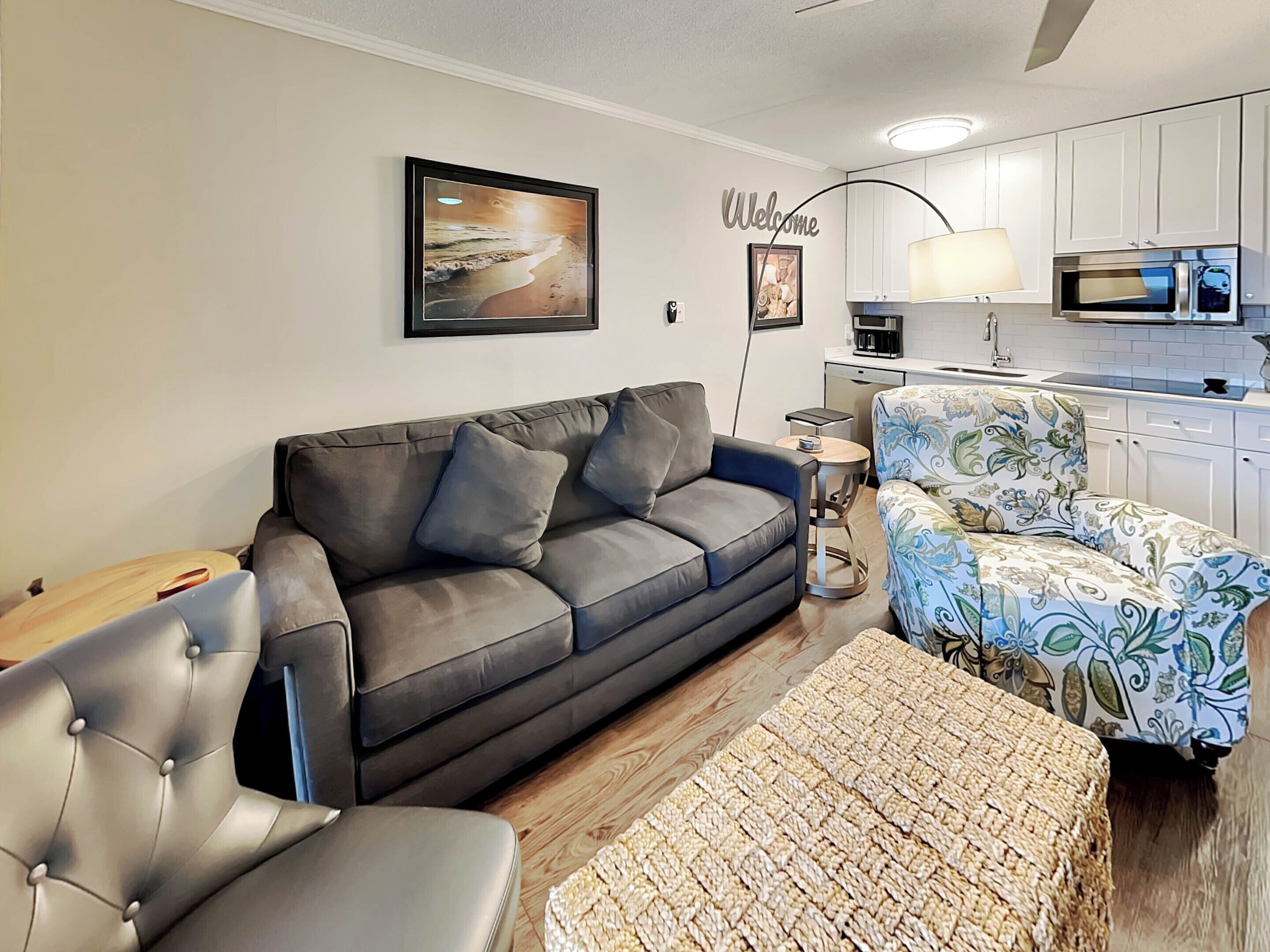 A living room with a grey sofa, a patterned armchair, a woven ottoman, a small side table, and a framed picture of a beach scene.