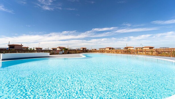 A large, clear swimming pool with a wooden deck surrounding it, set against a backdrop of buildings and a clear sky. - Villa Majanicho with pool (Majanicho)