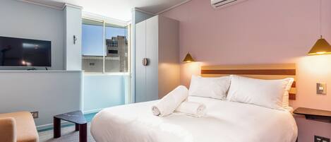 A modern hotel room with a large bed, a flat-screen TV, a small table, and a window with a view of buildings.