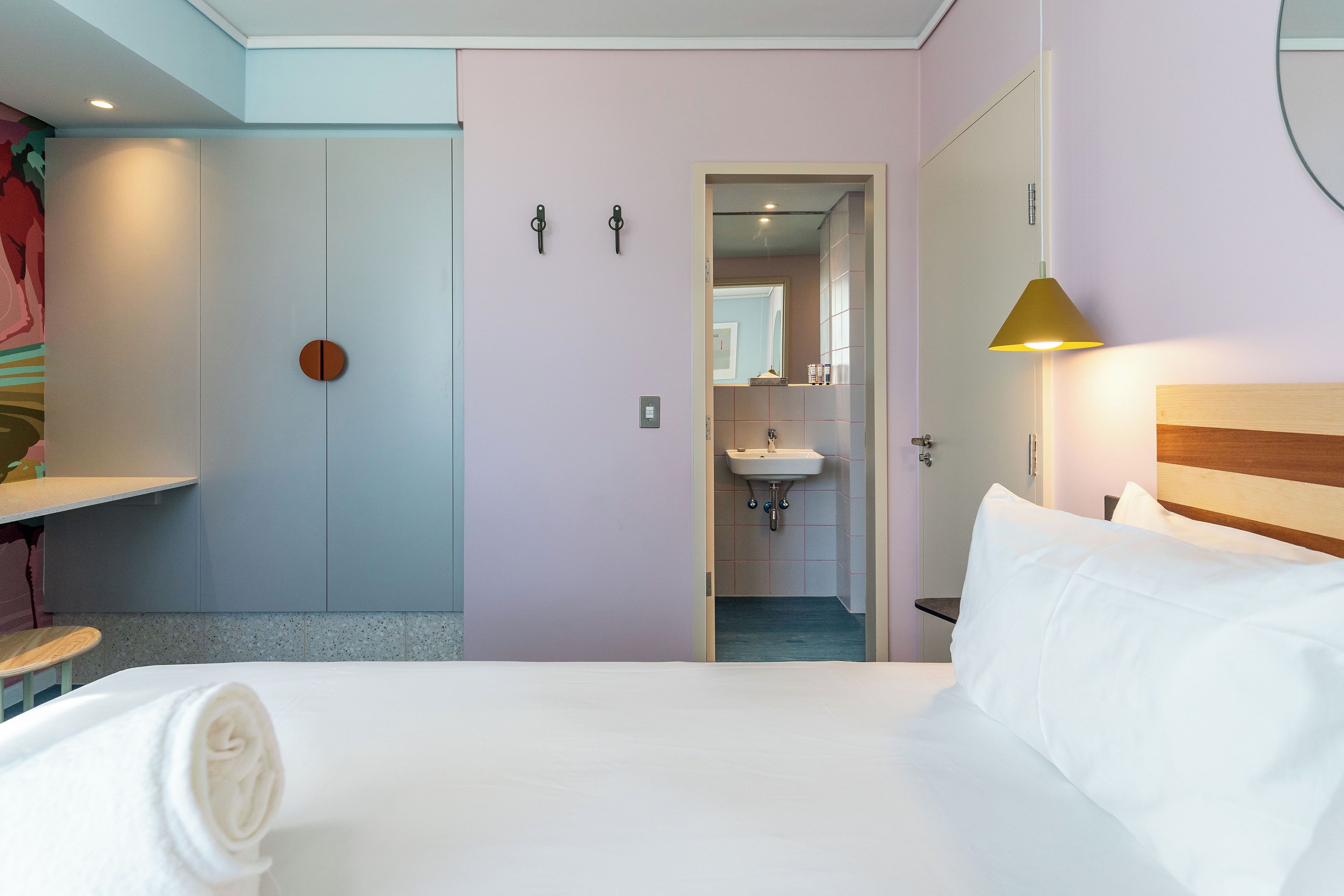 A hotel room with a bed, a bedside table, a lamp, a door leading to a bathroom, and a wall-mounted towel rack.
