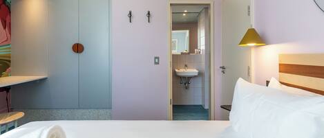 A hotel room with a bed, a bedside table, a lamp, a door leading to a bathroom, and a wall-mounted towel rack.