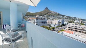 A balcony with white chairs and a table, overlooking a cityscape with buildings and a mountain.