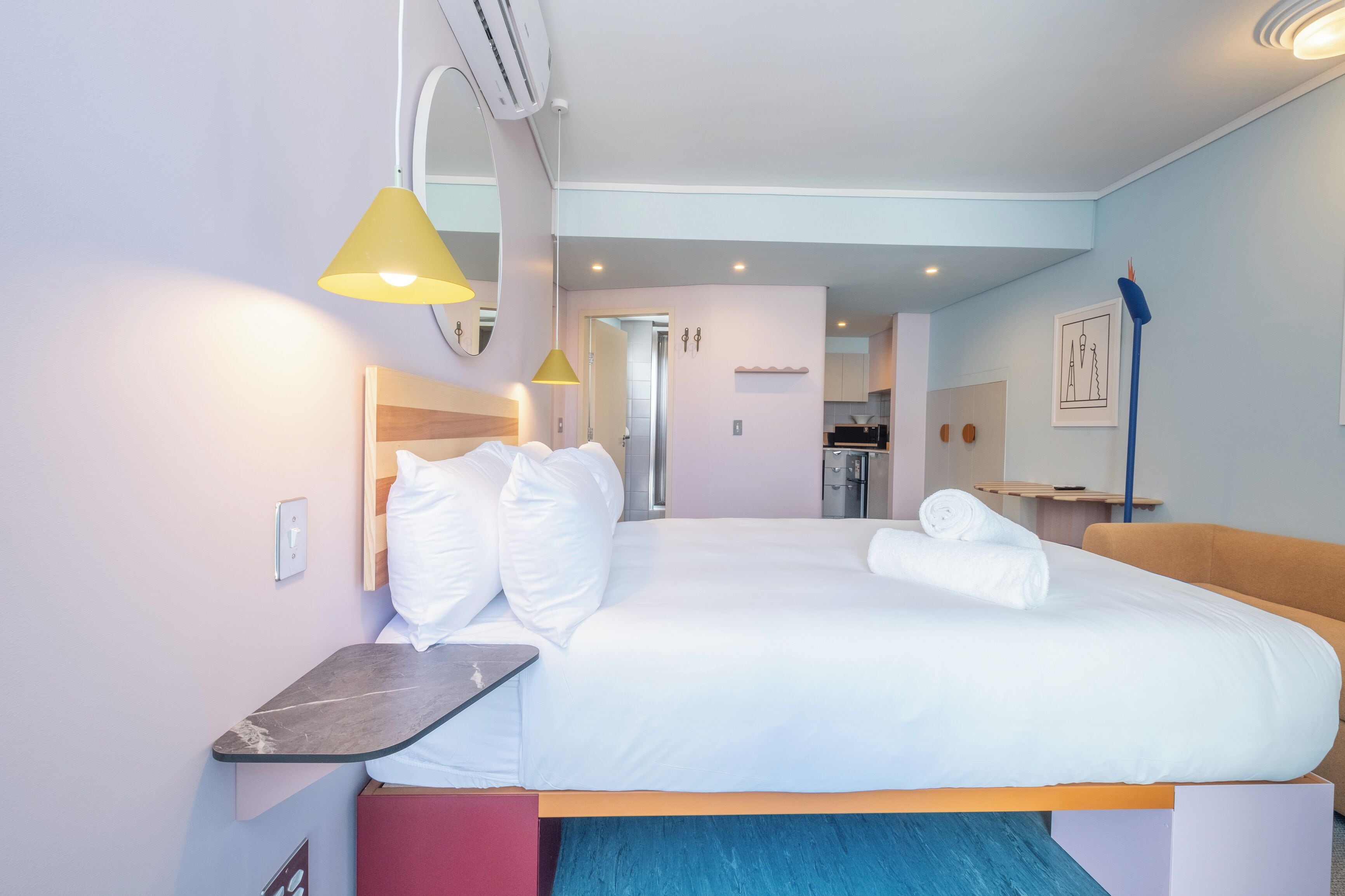 A hotel room with a bed, bedside table, and a small desk. There is a mirror above the bed, a wall-mounted shelf, and a door leading to a bathroom.