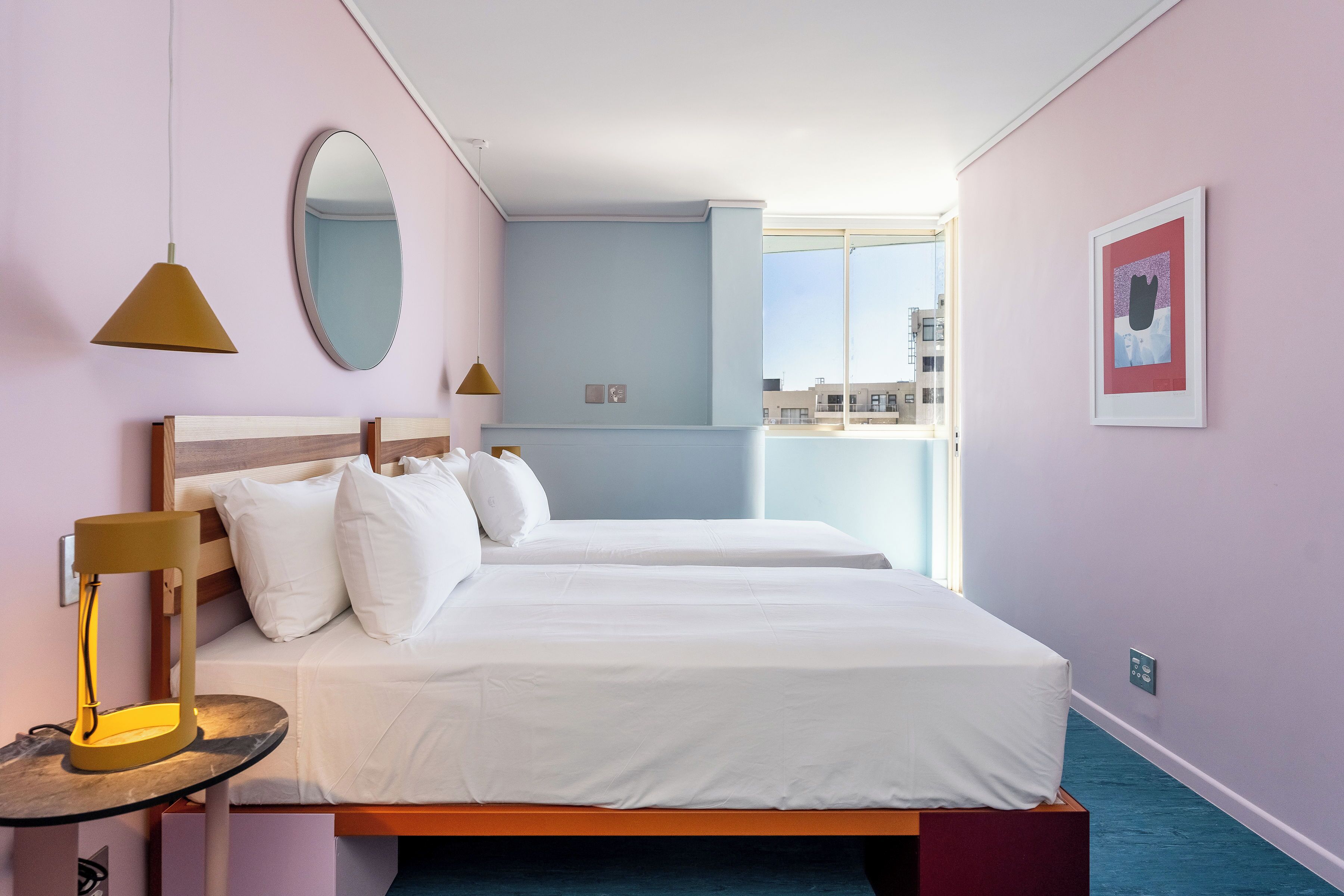 Habitación de hotel con dos camas, un espejo grande, una mesita de noche con lámpara y una ventana con vistas a la ciudad.
