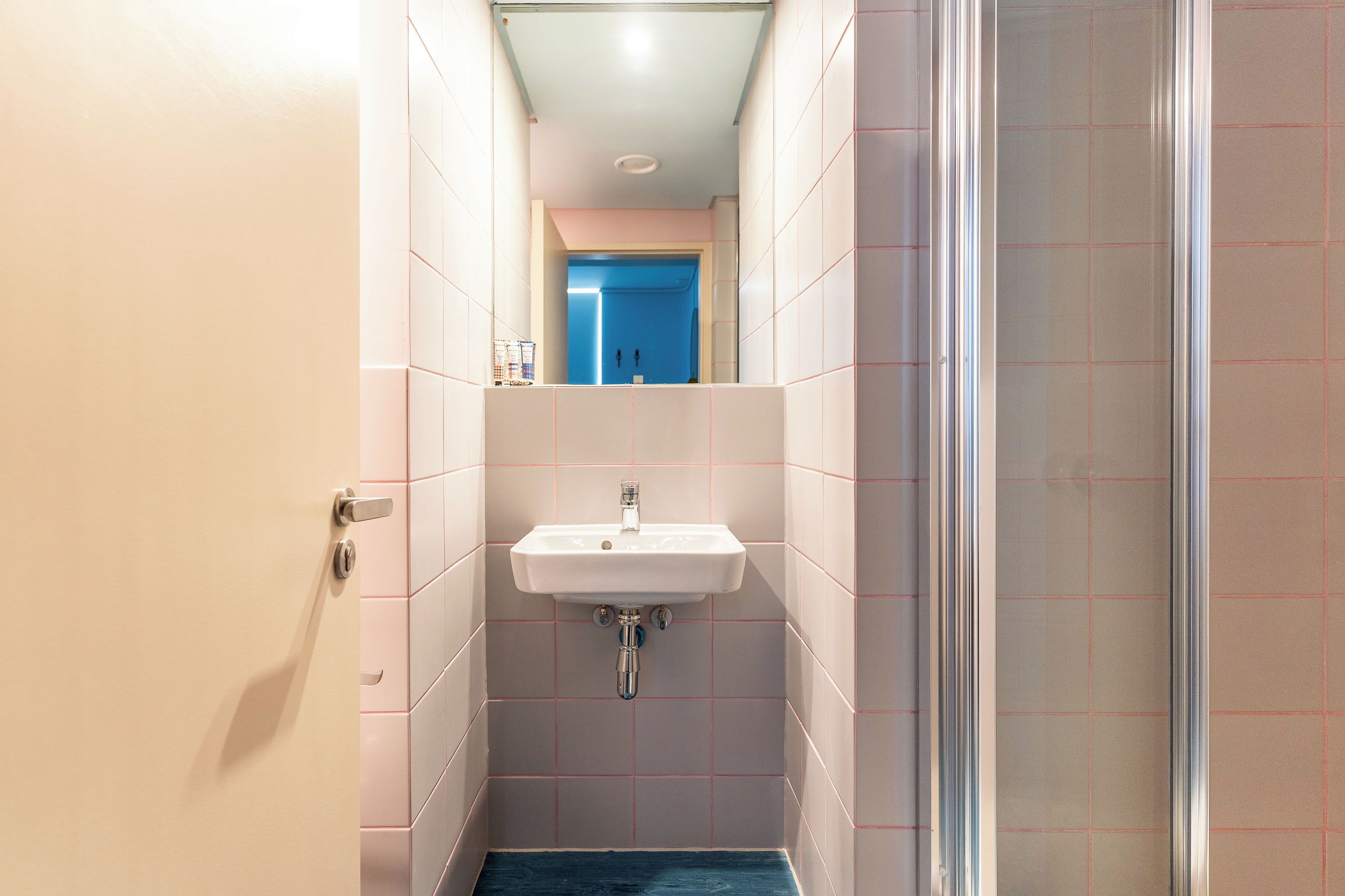 A narrow bathroom with a white sink, a blue shower area, and a door leading to another room.