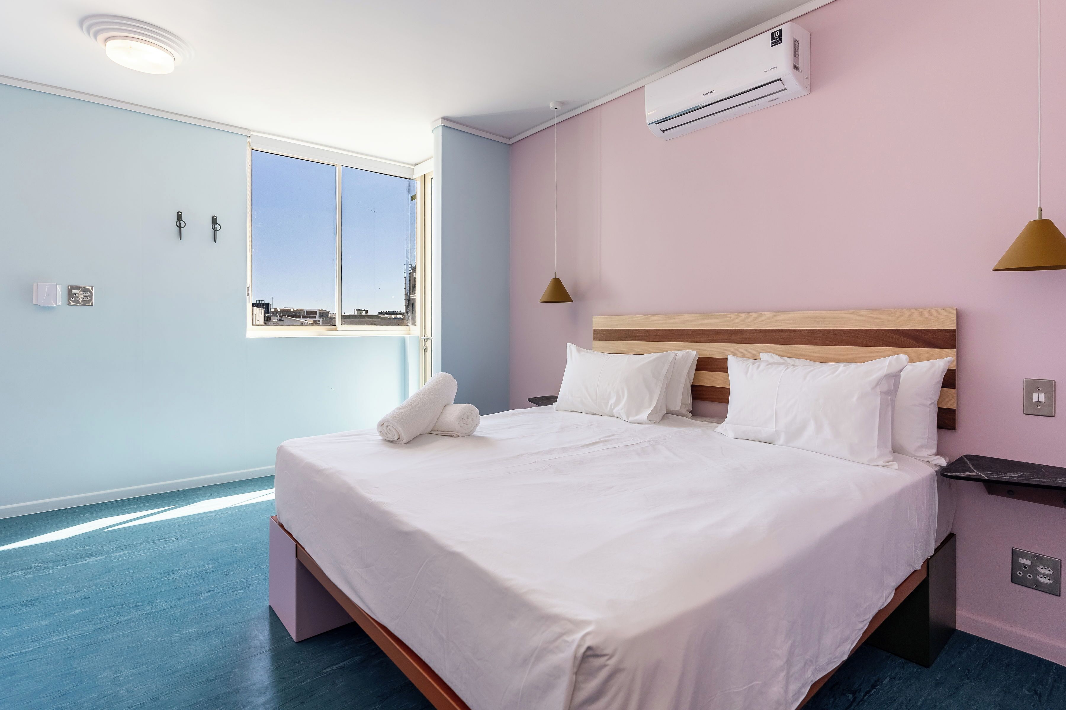 A neatly made bed with white linens and pillows, a window with a view, and an air conditioning unit on the wall.