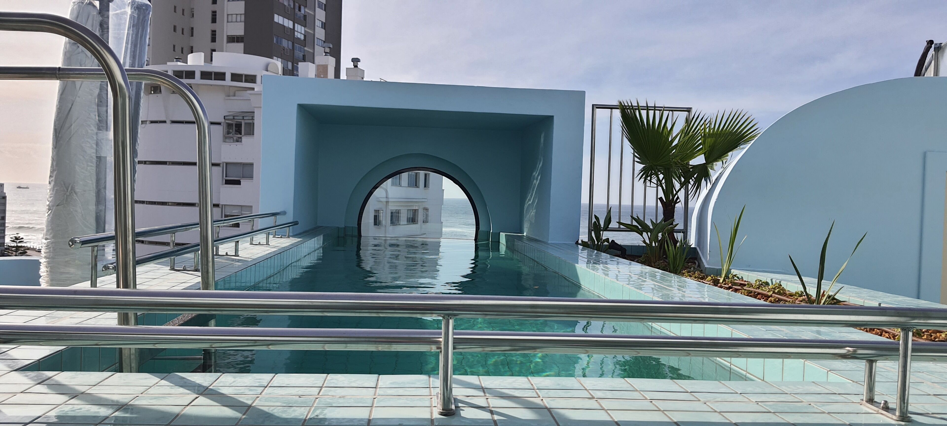 A swimming pool with a glass barrier, a blue archway, and a view of the ocean.