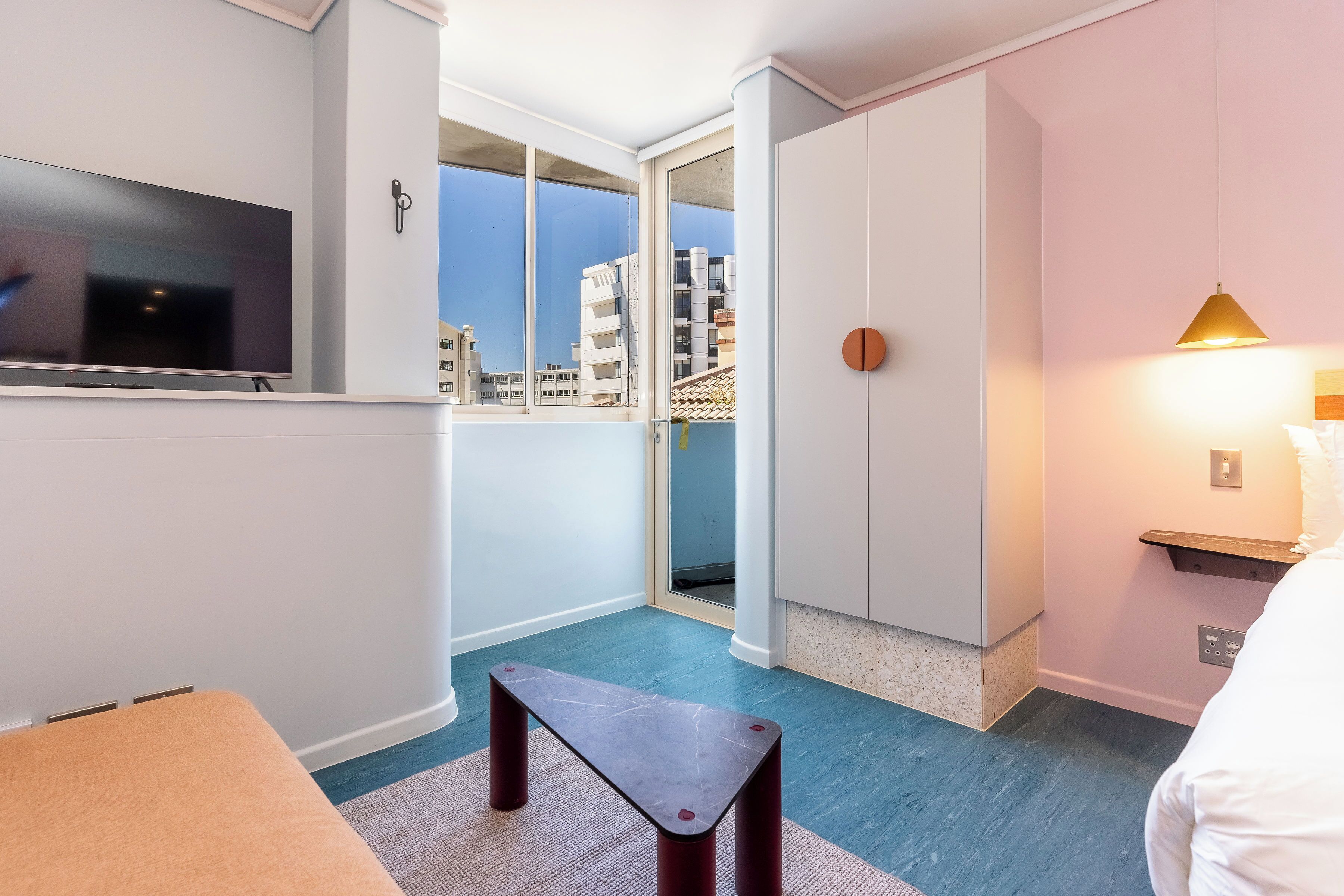 A hotel room with a large TV, a small table, a sofa, a balcony with a view of buildings, and a wall-mounted light fixture.