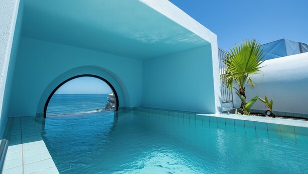 A clear blue pool with a view of the sea, a white building, and a palm tree.